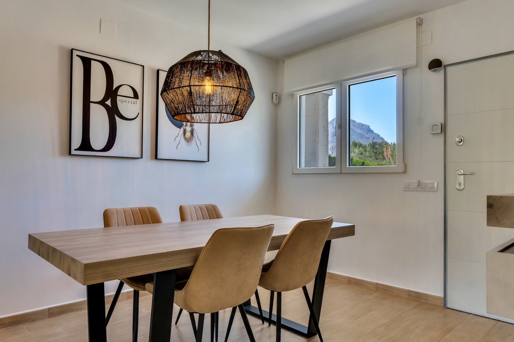 Modern dining area featuring a wooden table, upholstered chairs, and a woven pendant lamp. The window provides a clear view of the Montgó mountain.
