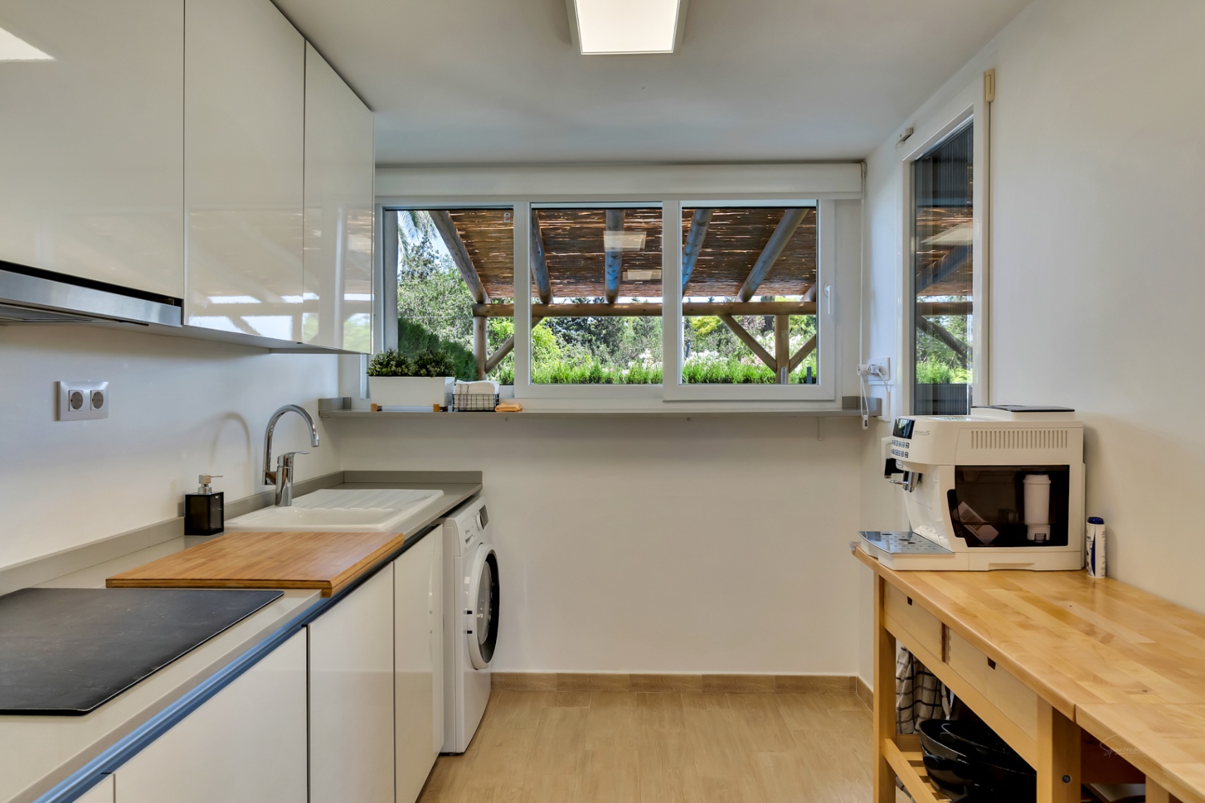 Modern kitchen featuring glossy white cabinets, integrated laundry area, and a large window overlooking the private patio's wooden pergola structure.
