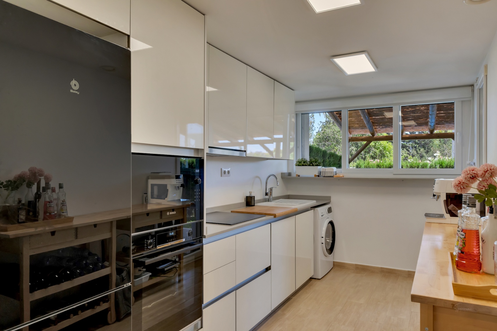 Modern kitchen featuring high-gloss white cabinetry, integrated appliances, and a large window overlooking the private patio and wooden pergola.