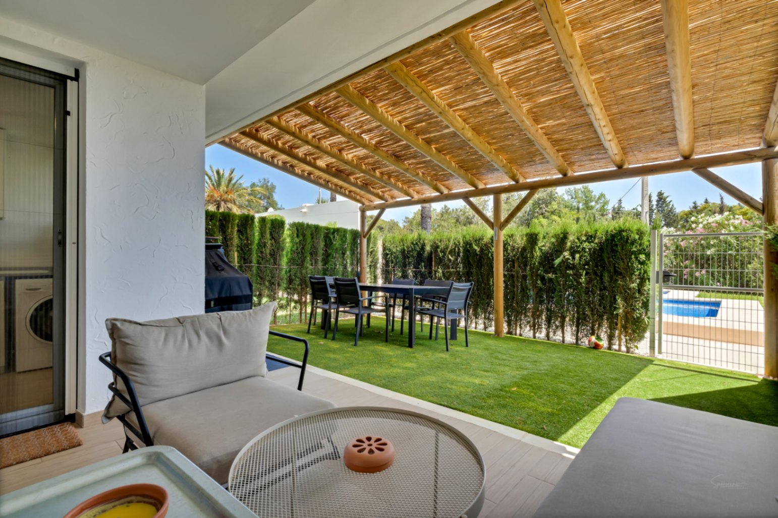 Private patio featuring a wooden pergola with reed roofing, outdoor dining area, and artificial turf. A secure gate provides direct access to the communal swimming pool.