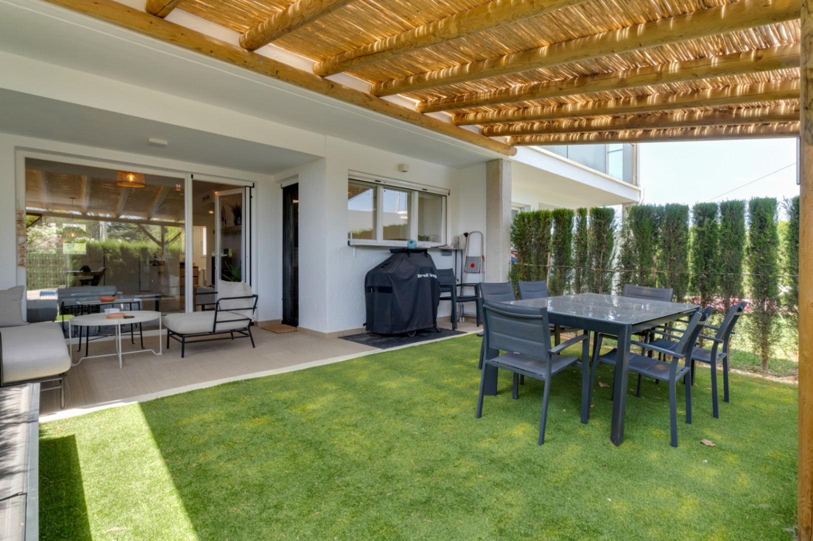 Private patio with artificial grass, a wooden pergola, and an outdoor dining area. It features a lounge set and barbecue, with direct access to the living room.