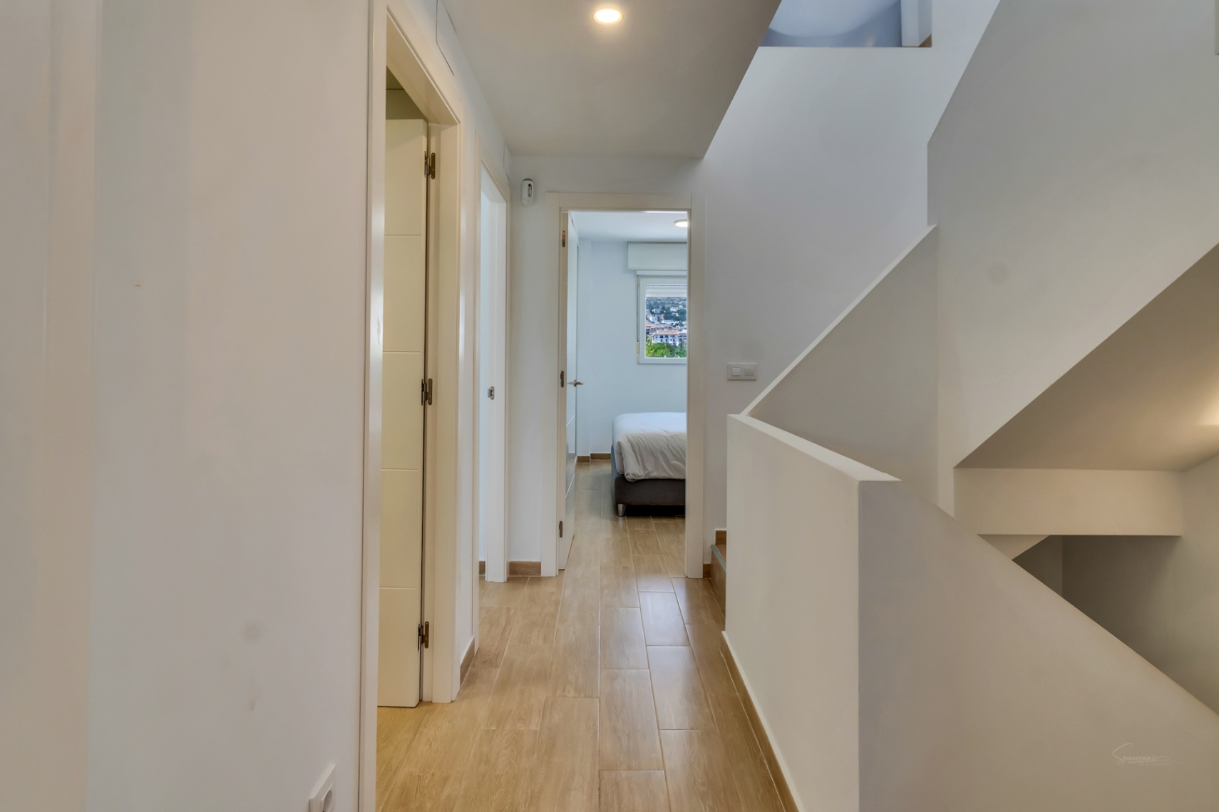Upper floor landing featuring wood-effect ceramic tiles and a minimalist white staircase. A view into a bright bedroom reveals natural light and town vistas.