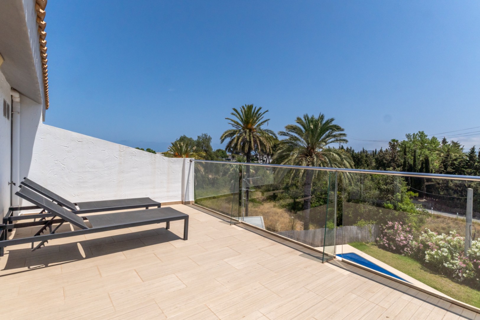 Upper-level terrace featuring ceramic tiling, glass balustrades, and sun loungers, overlooking palm trees and Mediterranean greenery.