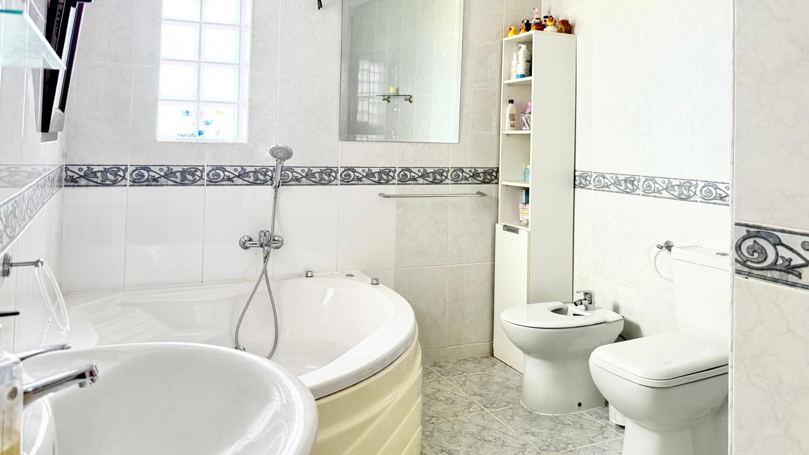 Bathroom featuring a corner whirlpool tub, bidet, and toilet. Finished with white wall tiles, a decorative border, and natural light from a glass block window.