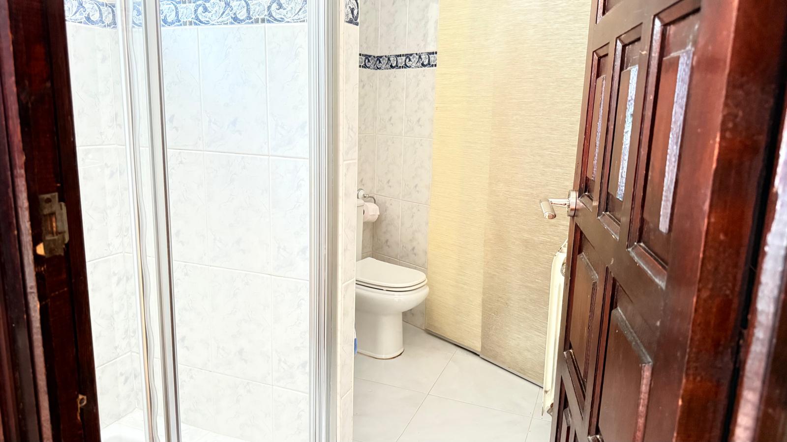 Bathroom featuring a glass-enclosed shower, toilet, and tiled walls with a blue decorative border. A dark wood door frames the foreground.