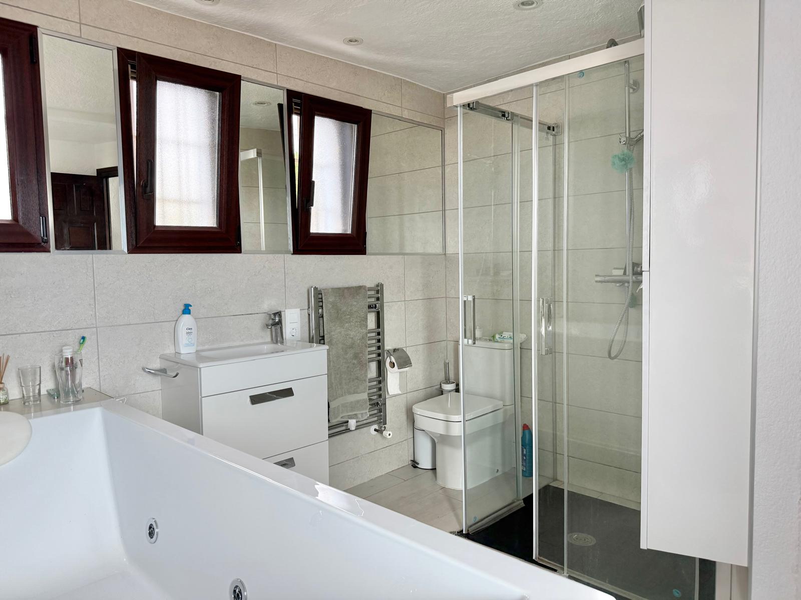 Bathroom featuring a hydro-massage bathtub, a separate glass-enclosed shower, a white vanity unit, and a heated towel rail set against neutral tiling.