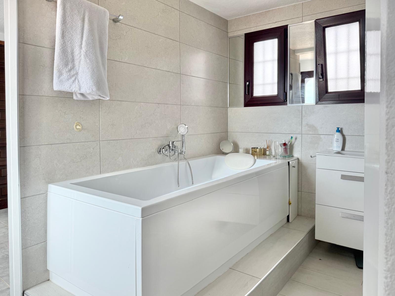 Bathroom featuring a white bathtub with headrest, neutral-toned large-format wall tiles, and a white vanity unit next to dark-framed windows.
