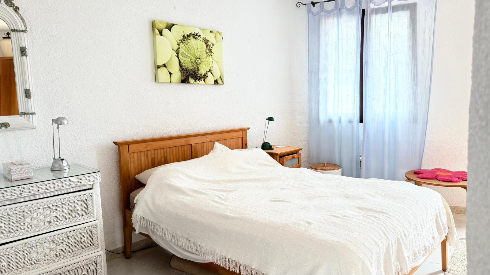 Bedroom featuring a wooden bed frame, white wicker chest of drawers, and textured walls. Natural light enters through a window with light blue translucent curtains.