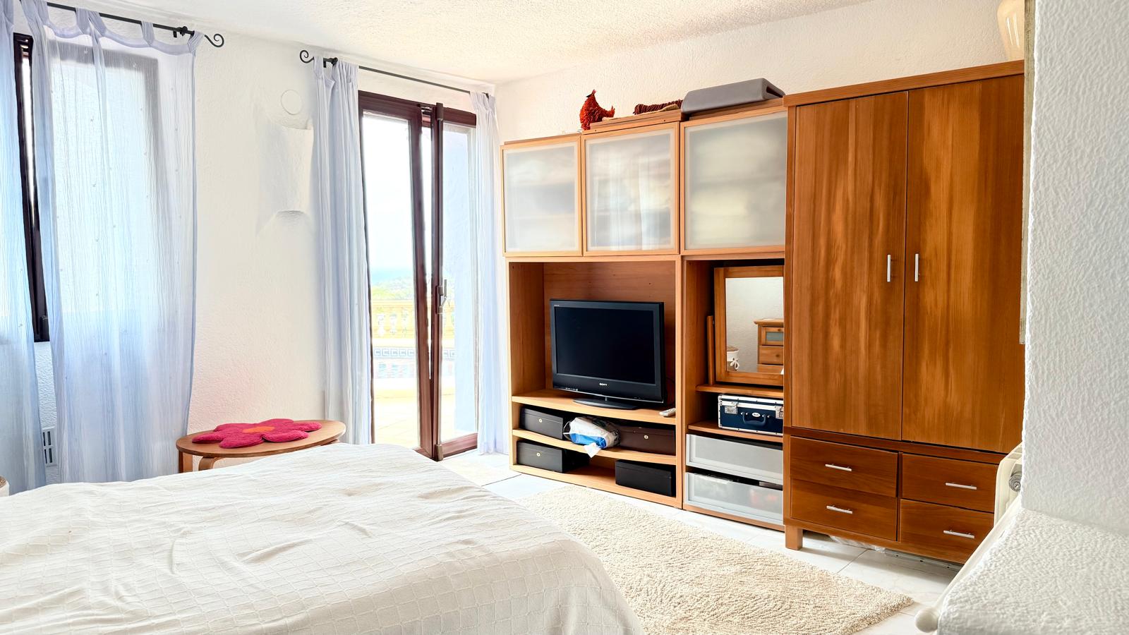 Bright bedroom featuring integrated wooden storage with a TV unit, a large wardrobe, and direct access to a terrace with coastal views.