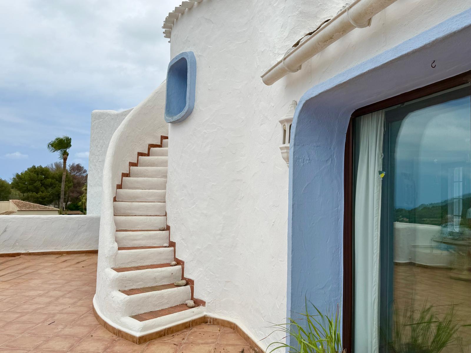 Curved Ibizan-style exterior staircase with terracotta treads, integrated into the white textured facade with blue-framed window accents.