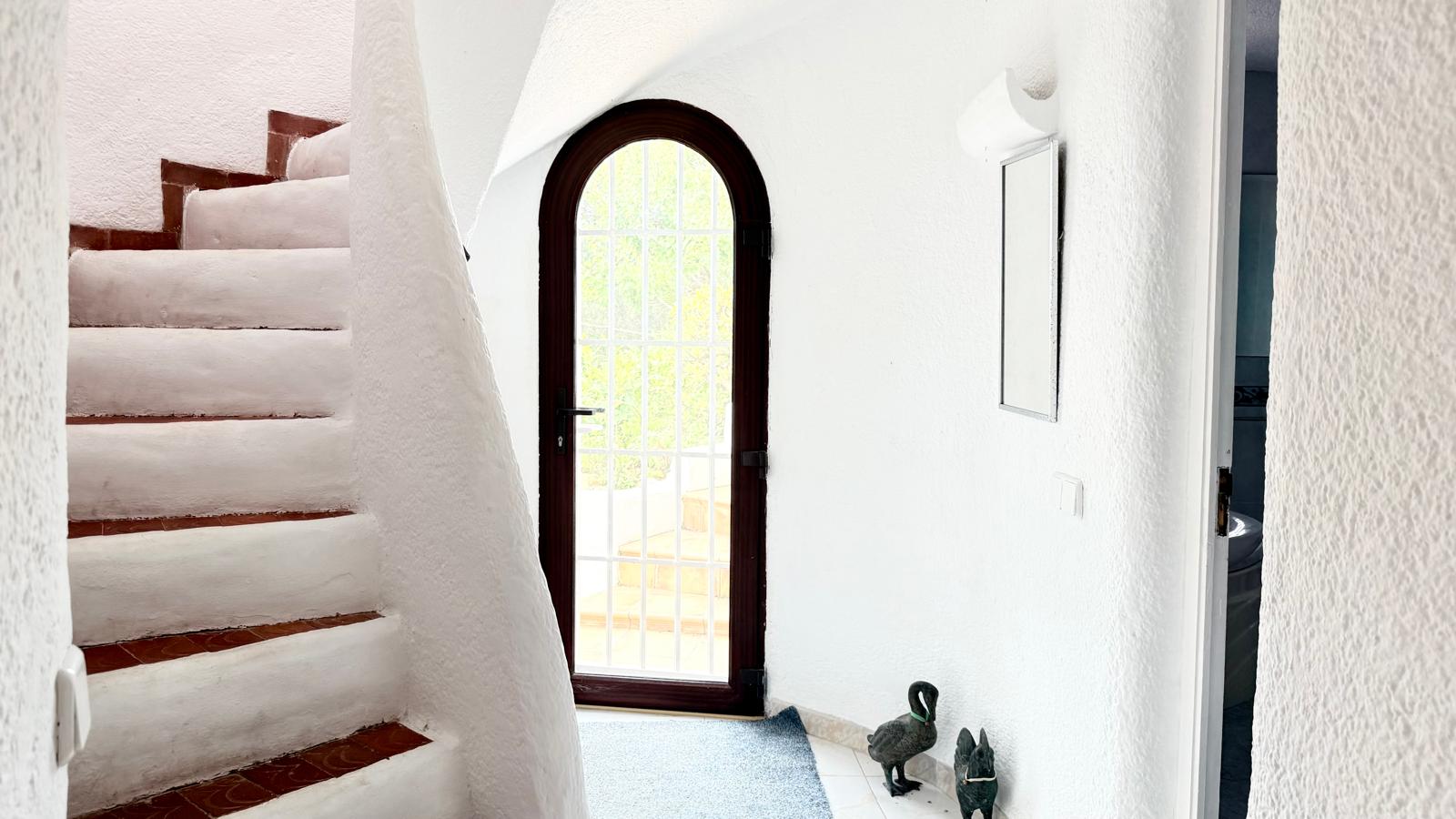 Curved Ibizan-style interior staircase featuring terracotta tile treads and textured white walls, adjacent to an arched glass door leading to the terrace.