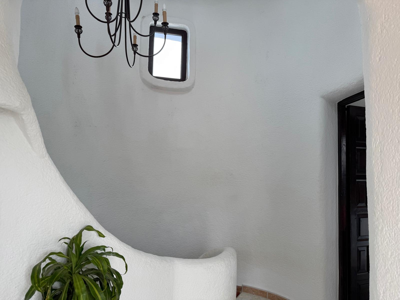 Curved Ibizan-style interior staircase featuring textured white plaster walls, a wrought-iron chandelier, and a small window providing natural light.