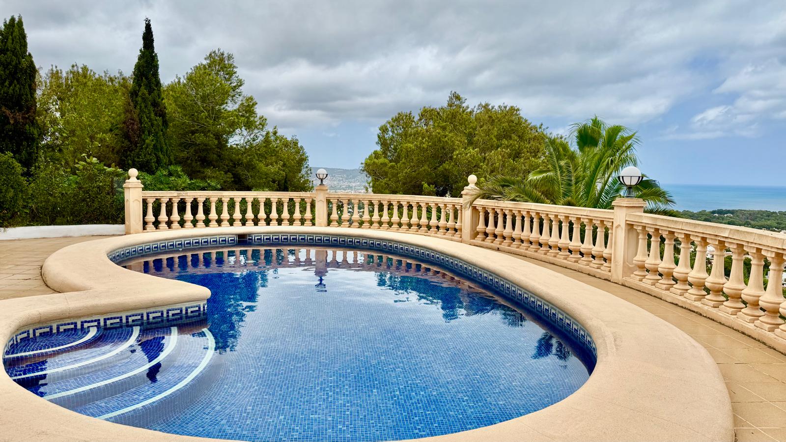 Curved private swimming pool featuring blue mosaic tiling, a stone-paved sun terrace, and a classic balustrade overlooking the Mediterranean Sea and pine trees.