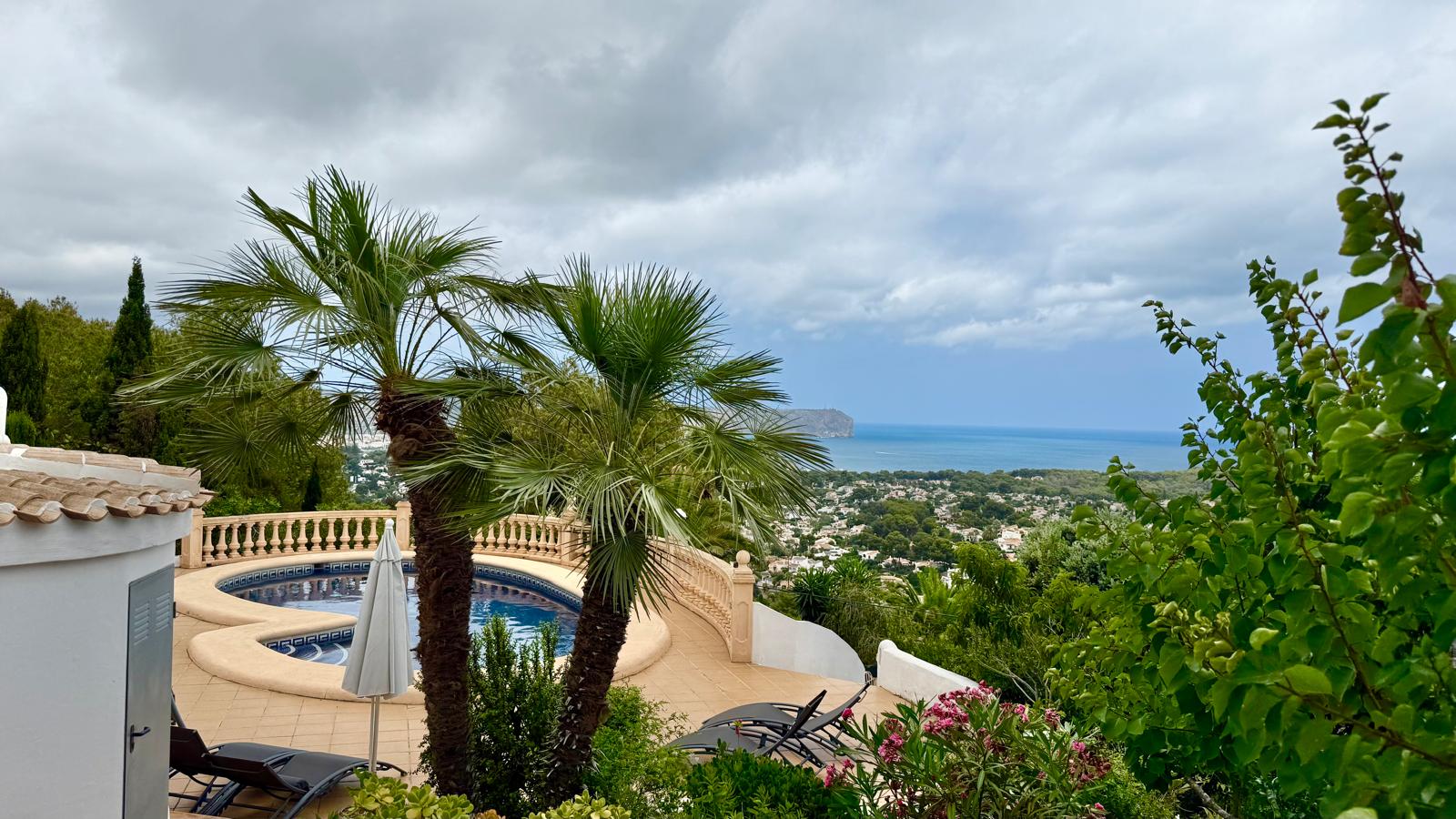 Elevated view of the curved swimming pool and Mediterranean gardens, offering a panoramic outlook over Jávea and the sea beyond mature palm trees.