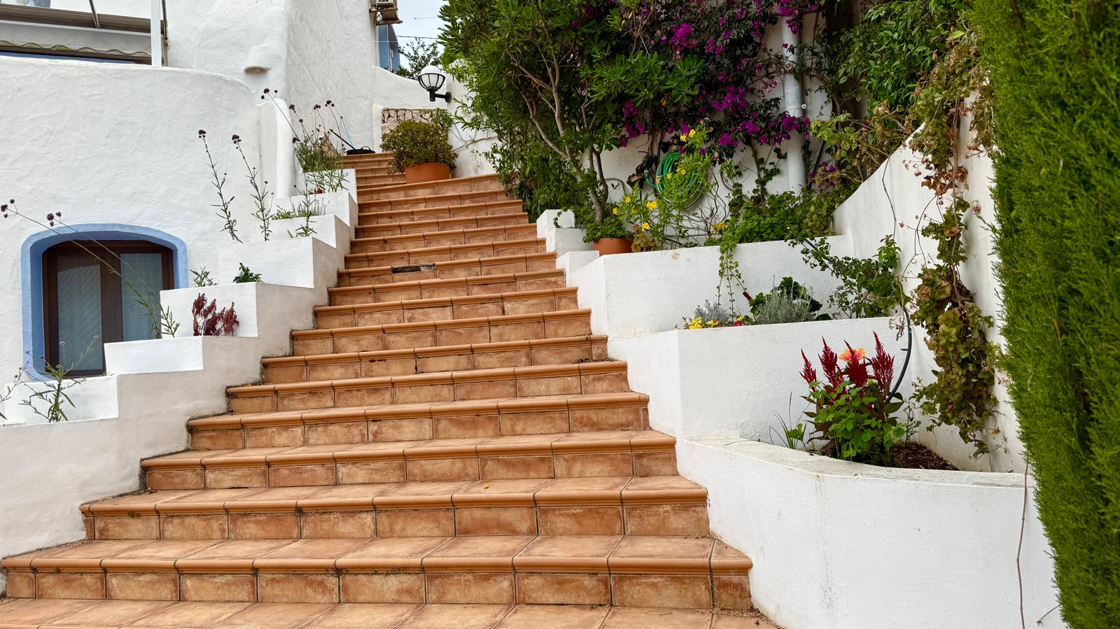 Exterior terracotta-tiled staircase connecting the villa's levels, bordered by white built-in planters with Mediterranean flora and vibrant bougainvillea.