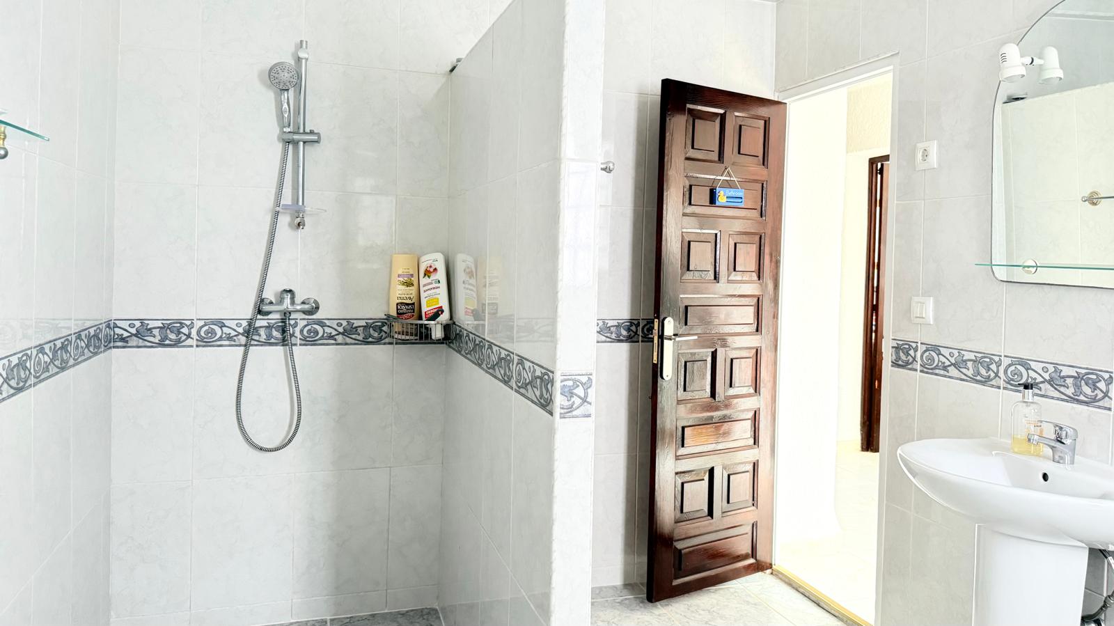Functional bathroom featuring a walk-in shower with decorative tile borders, a carved dark wood door, and a white pedestal sink with an illuminated mirror.