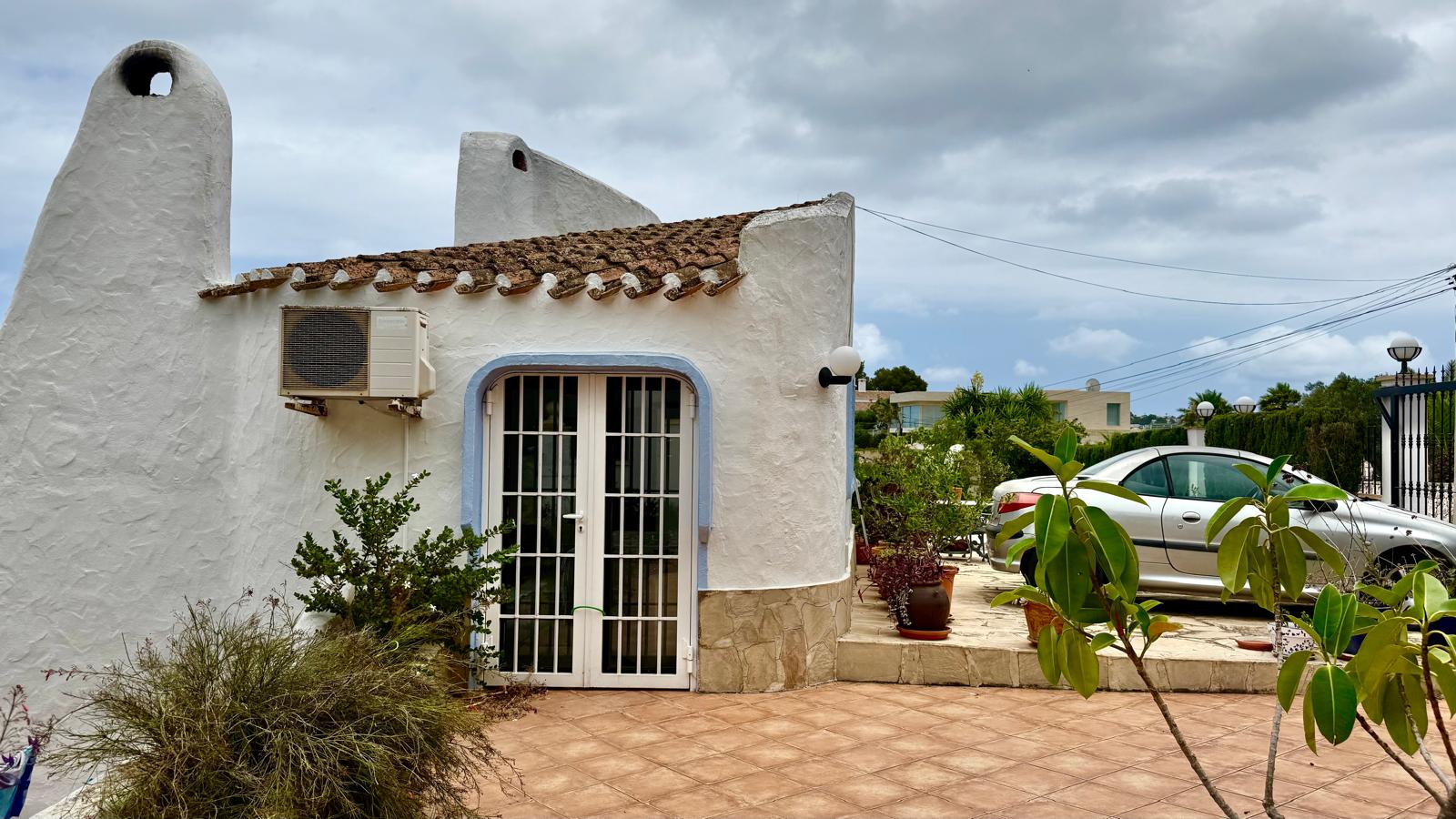 Ibicencan-style exterior featuring white-washed rounded walls, traditional chimneys, and a stone base. The level entrance includes a parking area and terracotta tiled terrace.