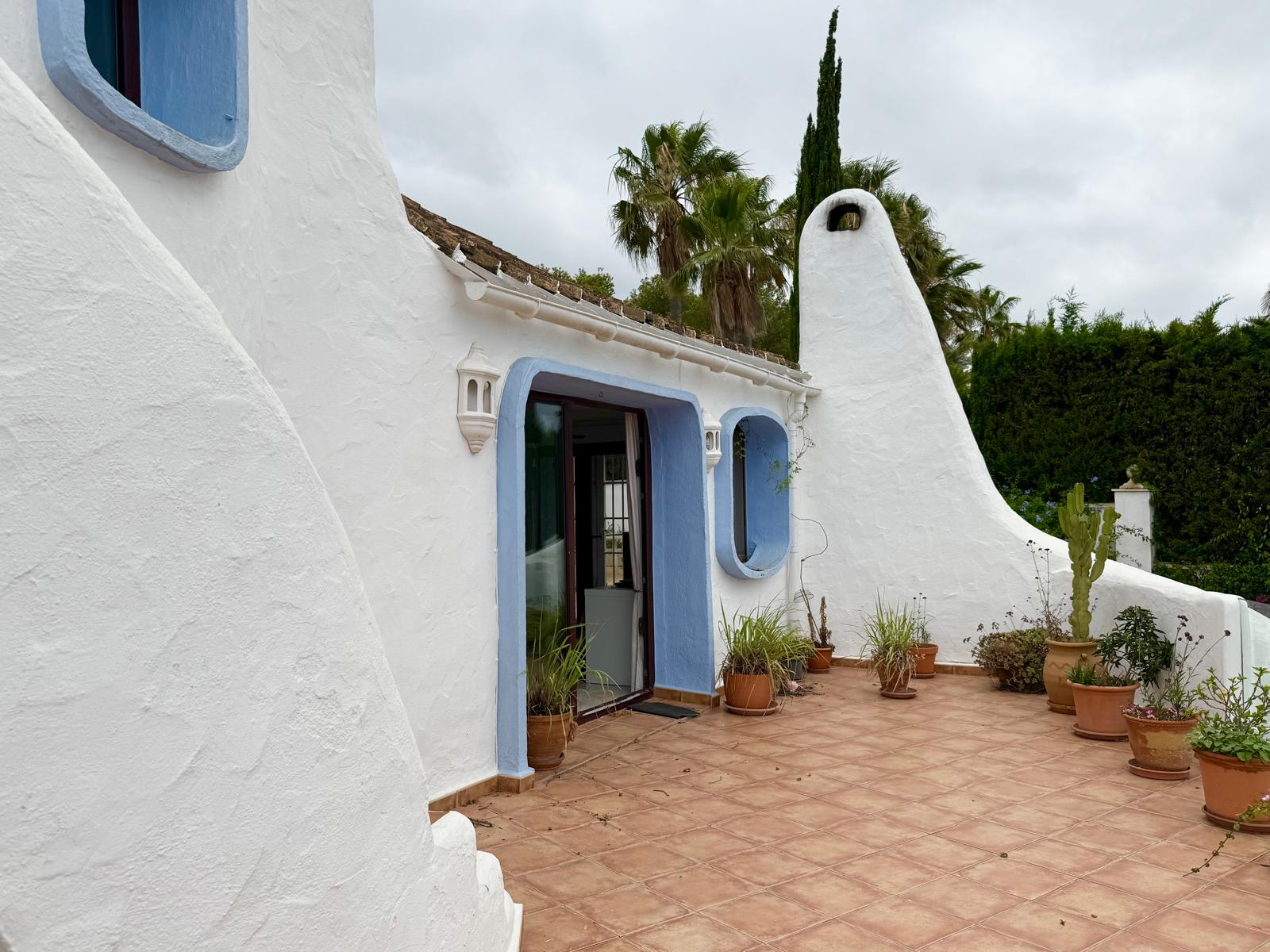 Ibicencan-style facade featuring organic white walls, blue-trimmed openings, and a sculptural chimney. The terracotta terrace is decorated with various Mediterranean potted plants.