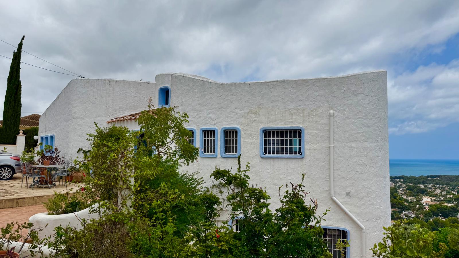 Ibicencan-style facade featuring textured white walls, blue window frames, and organic curves, set in an elevated position with panoramic Mediterranean views.