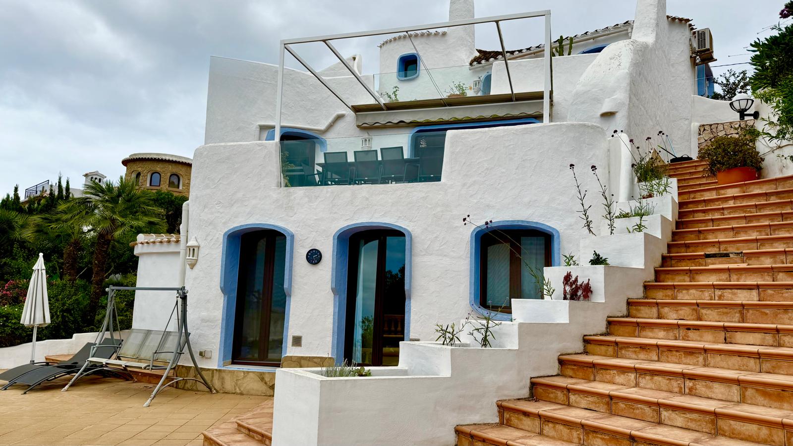 Ibicenco-style facade with white walls and blue trim. The photo shows the terraced levels connected by outdoor stairs, featuring a dining terrace and lower pool-level access.