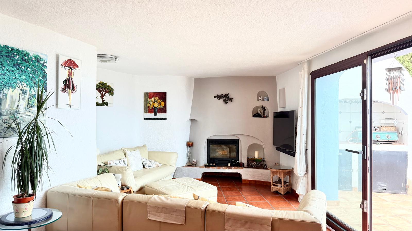 Ibicenco-style living room featuring a wood-burning fireplace, terracotta flooring, and curved walls. Large sliding glass doors provide direct access to the terrace.