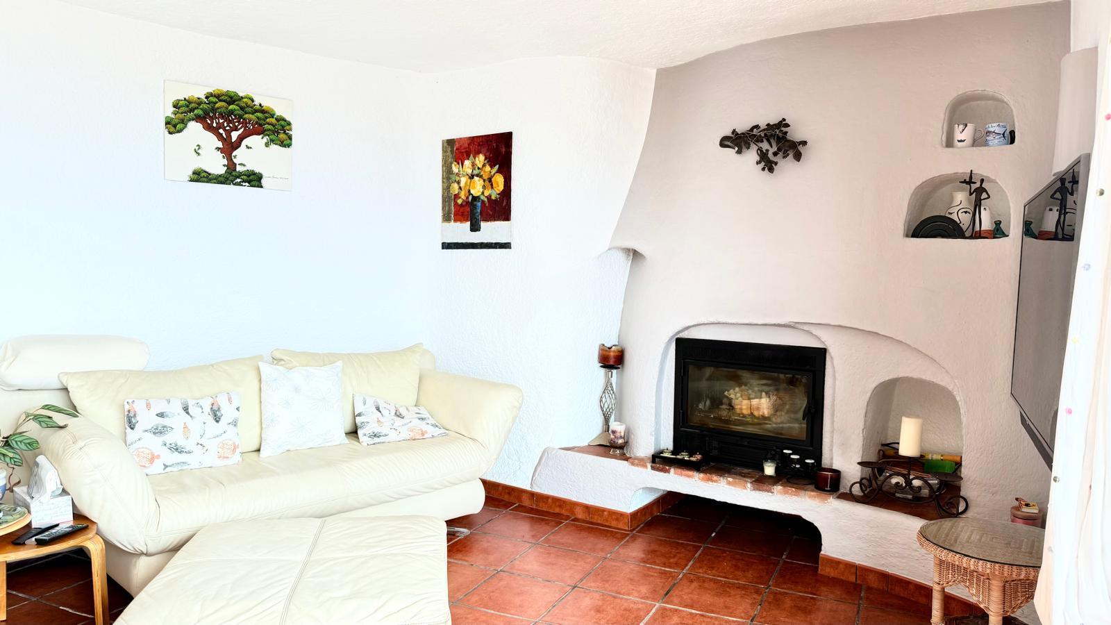 Ibiza-style living room featuring a wood-burning fireplace with a glass door, built-in wall niches, traditional terracotta flooring, and textured white walls.