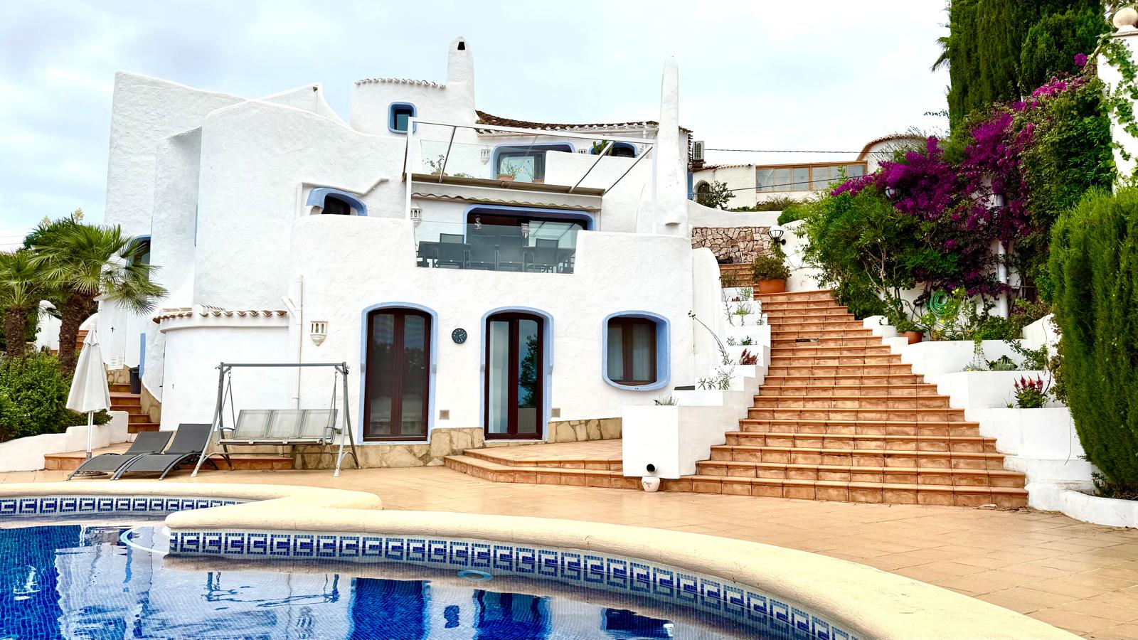 Ibiza-style villa featuring white curved walls, blue window trims, a large swimming pool, and terracotta stairs connecting the terraced Mediterranean gardens.