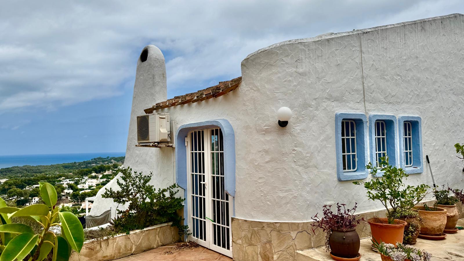 Ibizan-style exterior featuring curved white walls and blue-framed windows. The sculptural chimney and terracotta pots complement the panoramic Mediterranean views.