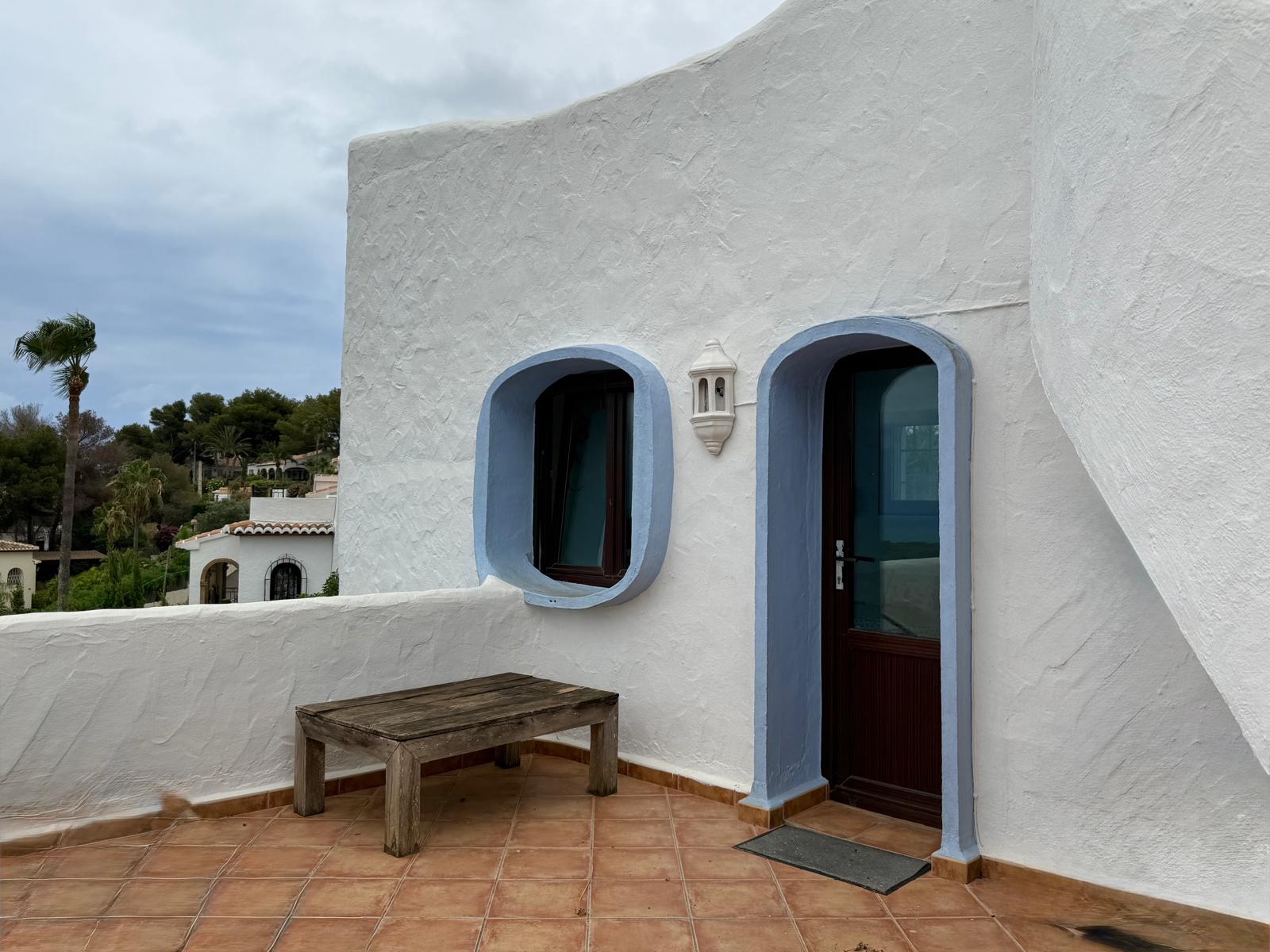 Ibizan-style facade featuring textured white walls, blue-trimmed arched openings, and terracotta flooring. The terrace includes a dark wood door and rustic outdoor furniture.