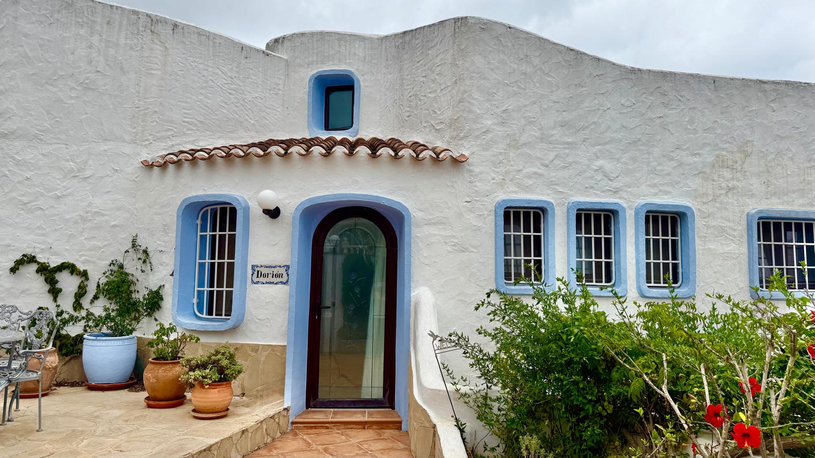 Ibizan-style facade featuring white textured walls, blue window trims, and an arched glass entrance door. The terrace shows terracotta tiling and Mediterranean potted plants.