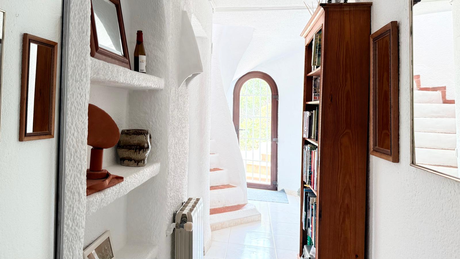 Ibizan-style hallway featuring textured white walls, built-in masonry shelving, and a curved staircase with terracotta accents leading toward an arched glass door.