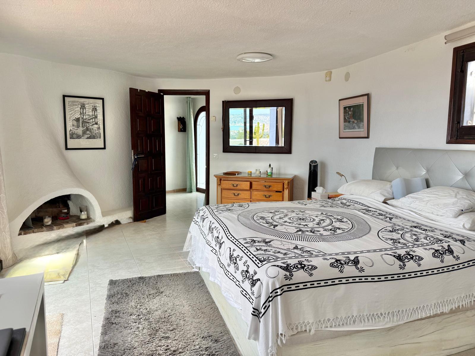 Ibizan-style master bedroom featuring a unique curved built-in fireplace, tiled flooring, and a mirror reflecting the panoramic sea views.
