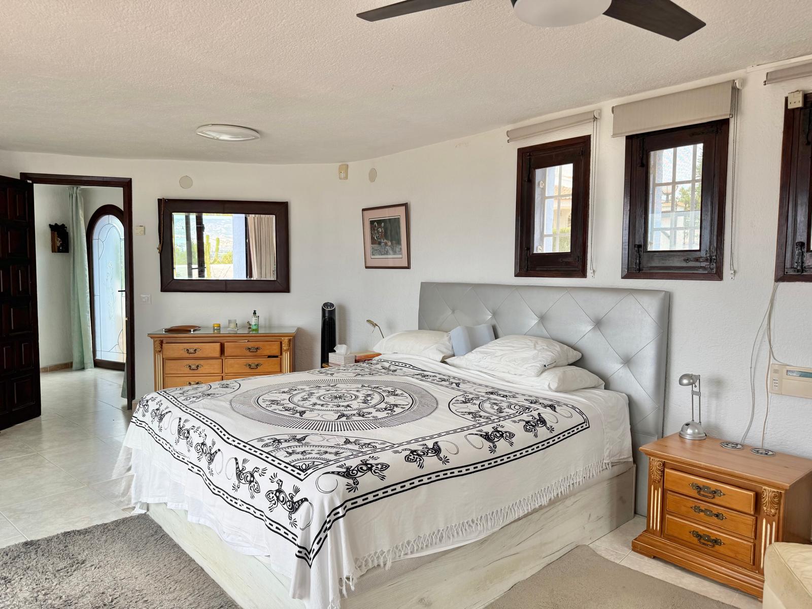 Master bedroom featuring white walls, dark wood window frames, and a ceiling fan. It includes classic wooden furniture and provides access to the adjacent dressing area.