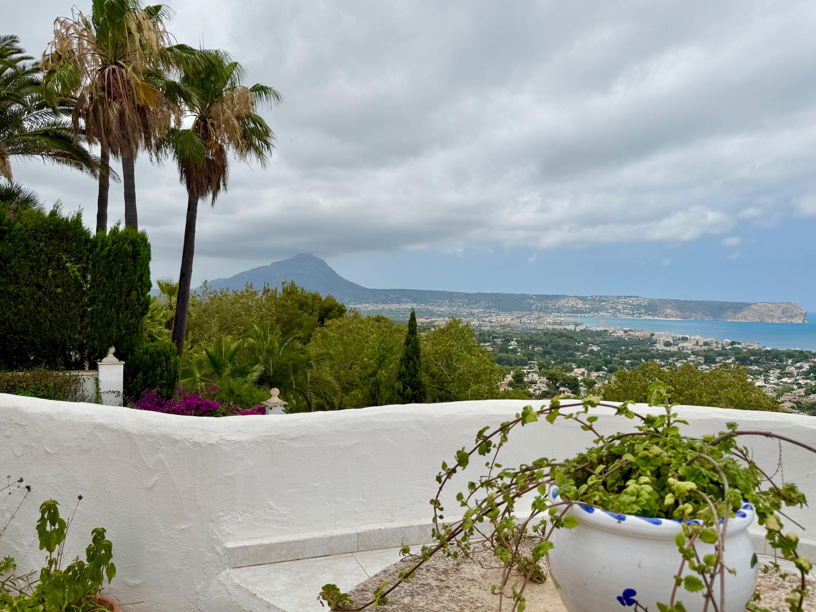 Panoramic view from the upper terrace overlooking Jávea Bay and Montgó mountain, framed by white Ibizan-style walls and mature Mediterranean greenery.