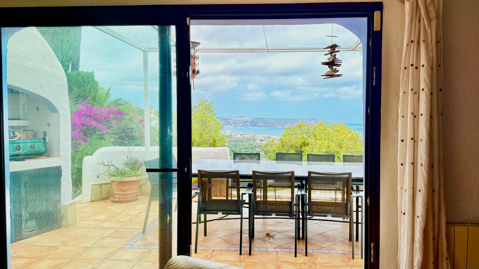 View from the living area looking out to the main terrace, featuring an outdoor dining set, built-in barbecue, and panoramic vistas of Jávea bay and the sea.