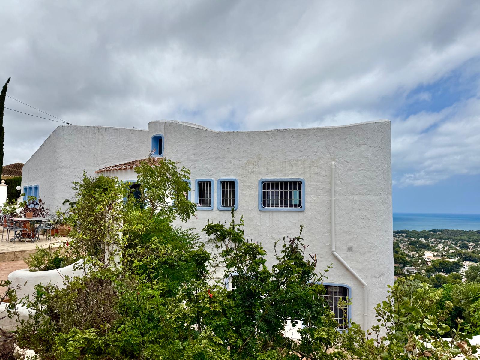 White Ibicenco-style facade featuring blue-trimmed windows and organic curved walls, overlooking the Mediterranean Sea and coastal landscape.