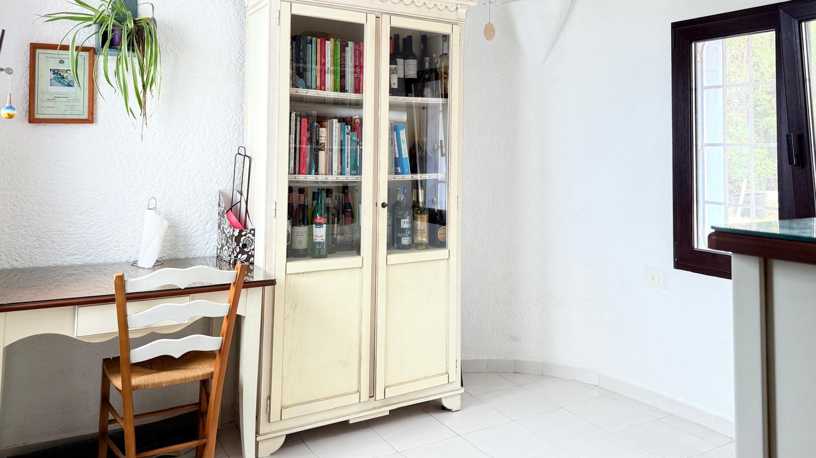Workspace featuring a white wooden desk, rustic chair, and a classic glass-fronted cabinet for books and bottles against textured white walls.