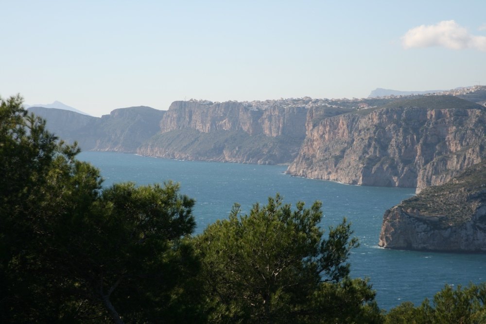 Panoramic views of the Mediterranean Sea and the rugged cliffs of the Jávea coastline, framed by native pine trees and a clear blue sky.