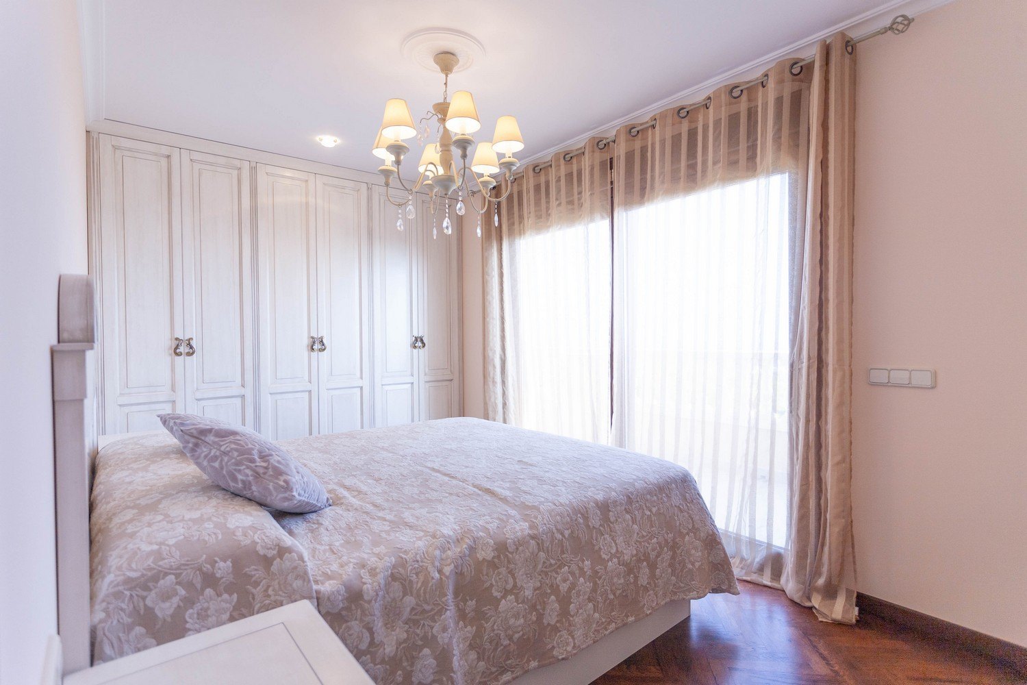 Bedroom featuring a double bed, white built-in wardrobe, chandelier, and beige curtains.