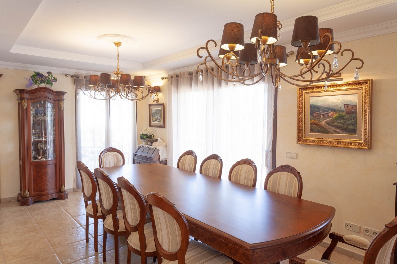 Classic dining room with wood table, upholstered chairs, and antique display cabinet. Warm lighting with chandeliers.