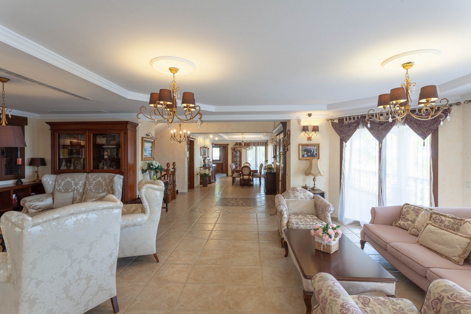 Open-plan living room featuring upholstered seating, bronze chandeliers, and tiled flooring.