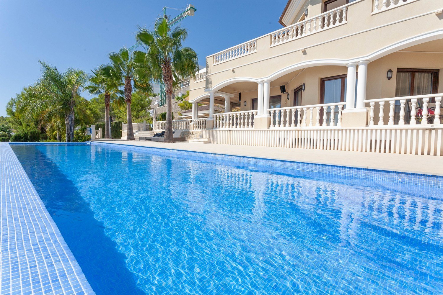View of the turquoise blue pool with mosaic border and the facade of the Mediterranean-style villa.