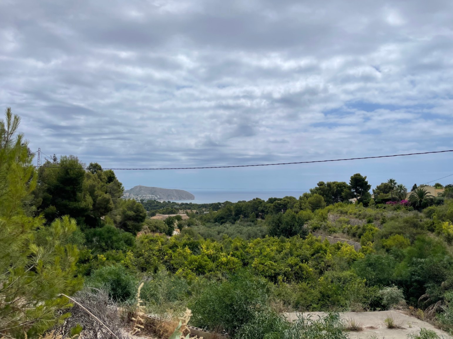 Panoramic view from the plot towards the Moraira coastline, featuring the Cap d'Or headland and the Mediterranean Sea framed by pine trees.