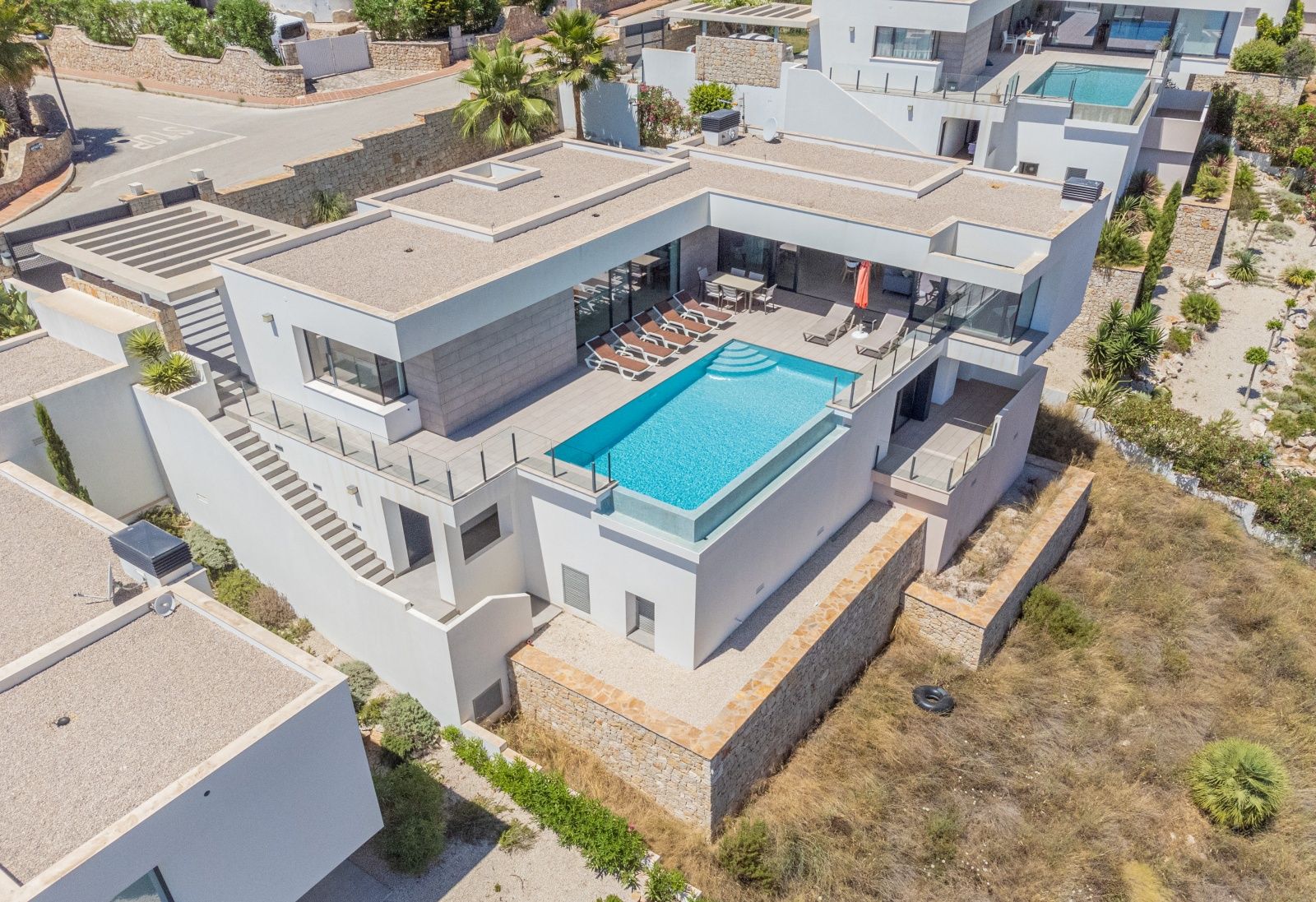 Aerial view of the modern villa featuring an infinity pool with glass wall, spacious sun terrace with loungers, and outdoor dining area integrated into the white architecture.