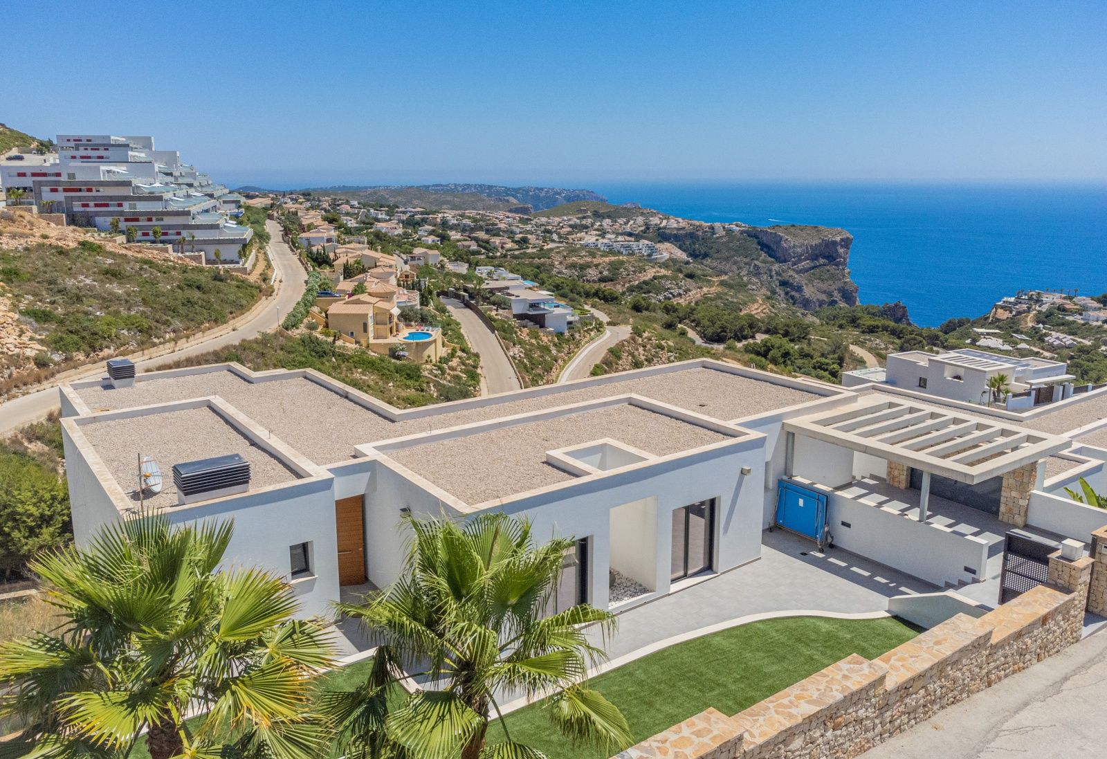 Aerial view of the modern villa in Cumbre del Sol, featuring white minimalist architecture, gravel flat roofs, and panoramic views of the Mediterranean coastline and cliffs.