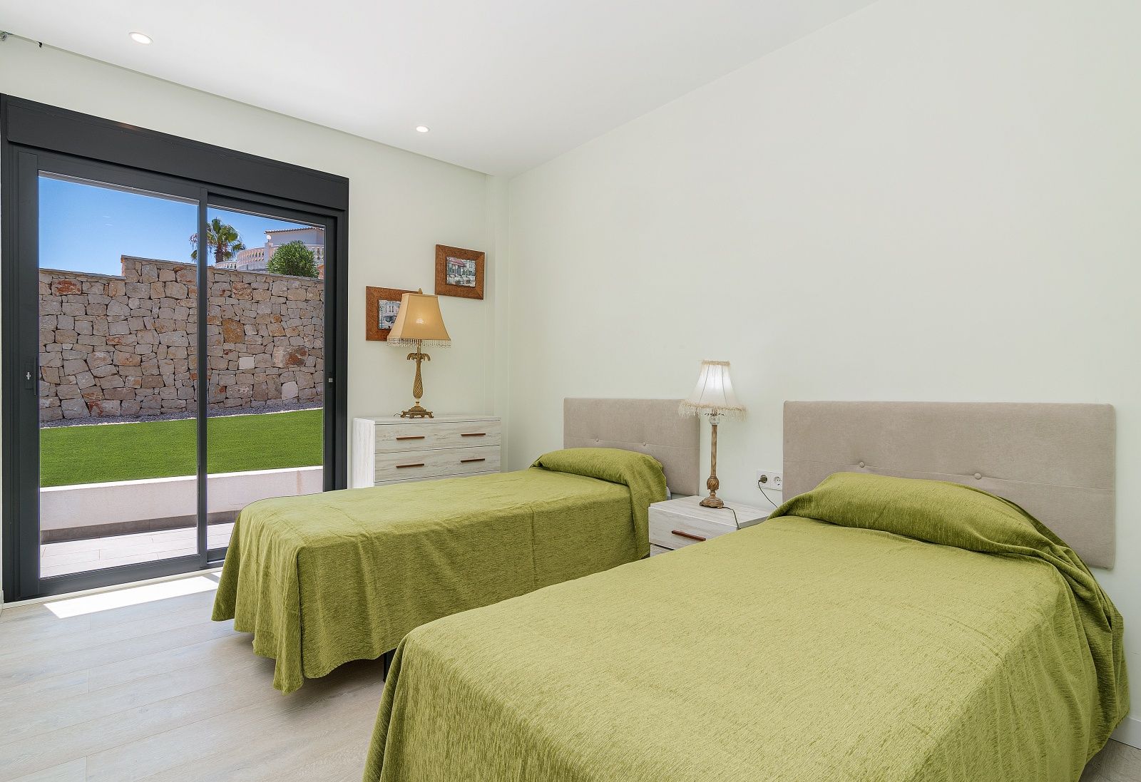 Bedroom featuring twin beds with upholstered headboards, recessed lighting, and large sliding doors opening to a terrace with a dry stone wall and lawn.