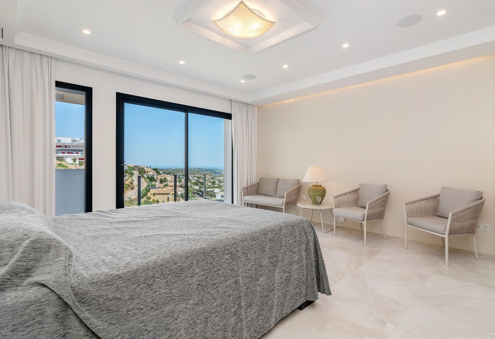 Bright bedroom featuring porcelain tile flooring, a seating area with modern armchairs, and floor-to-ceiling glass doors opening to sea and coastal views.