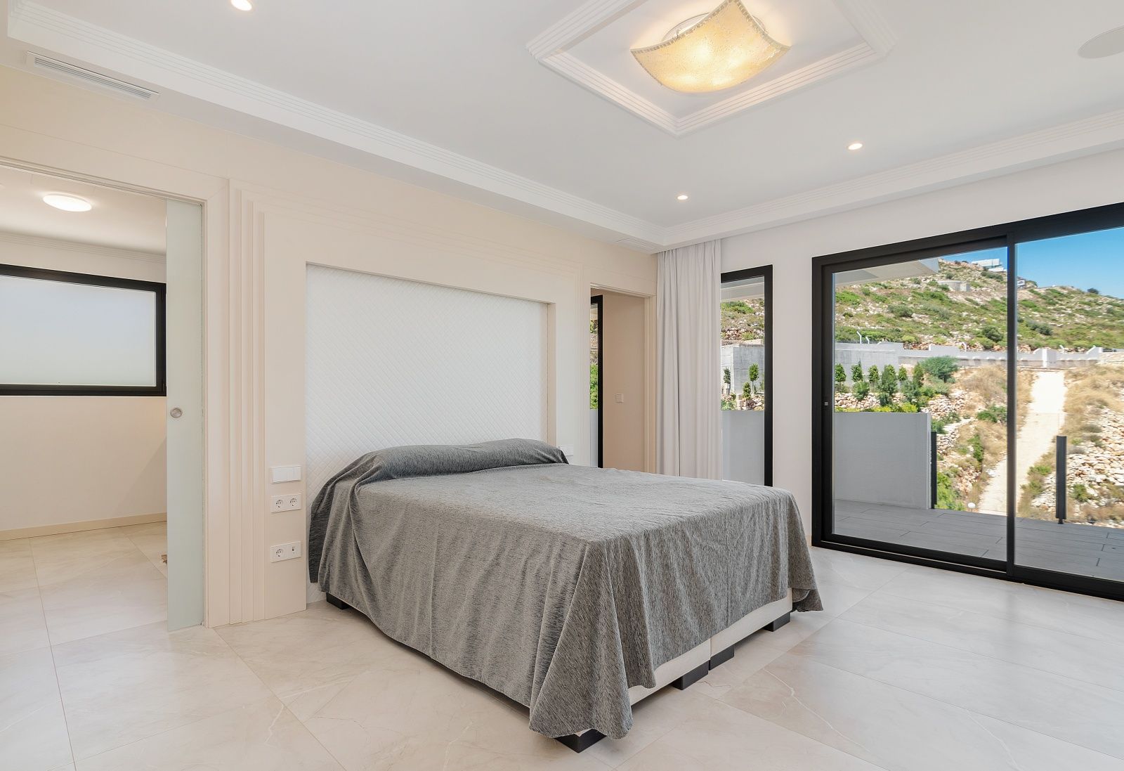 Bright bedroom featuring sliding glass doors to a terrace, recessed ceiling lights, and a textured headboard niche with elegant crown molding.