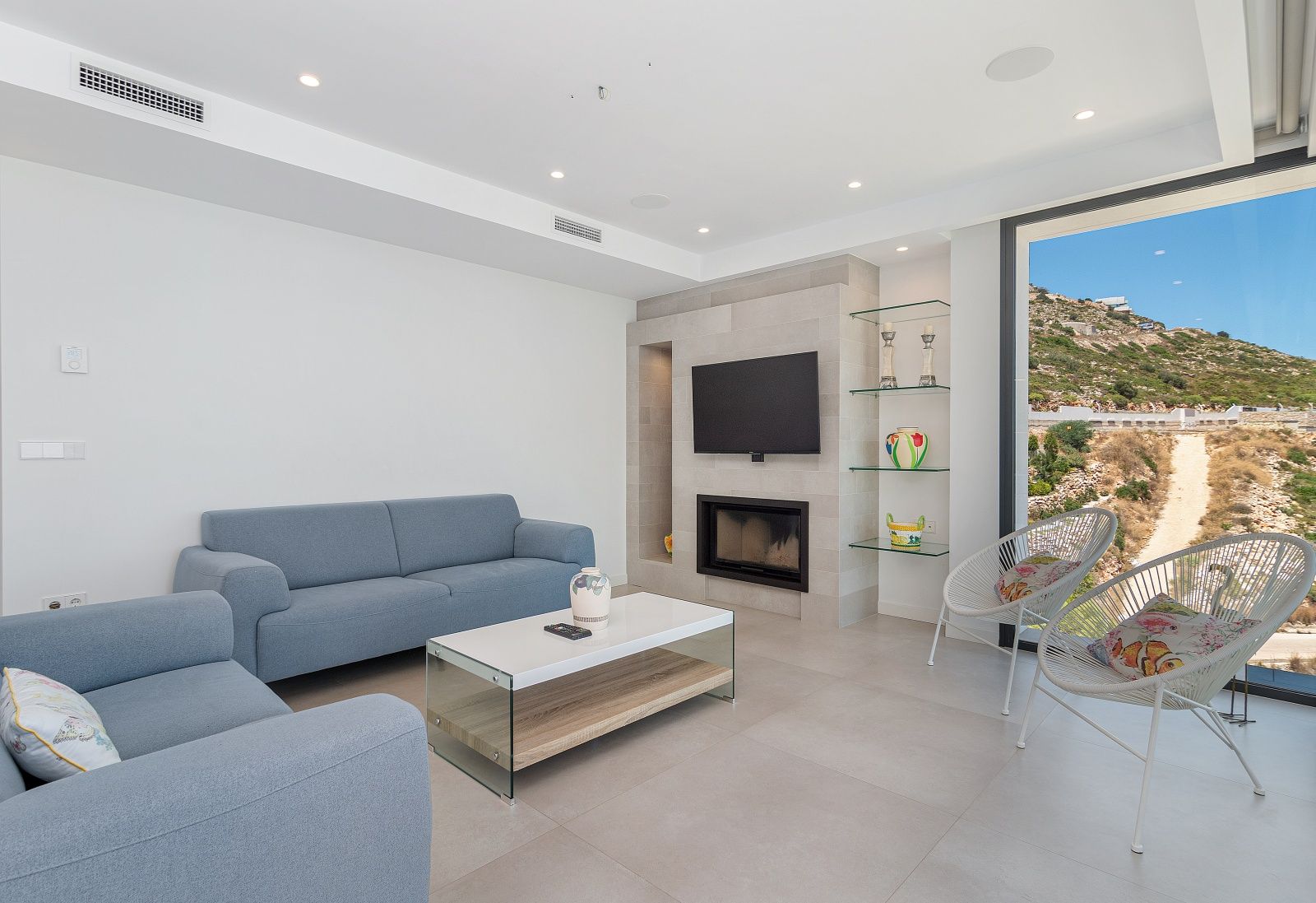 Contemporary living room featuring blue sofas, a stone-clad fireplace wall with integrated TV, and floor-to-ceiling windows overlooking the landscape.