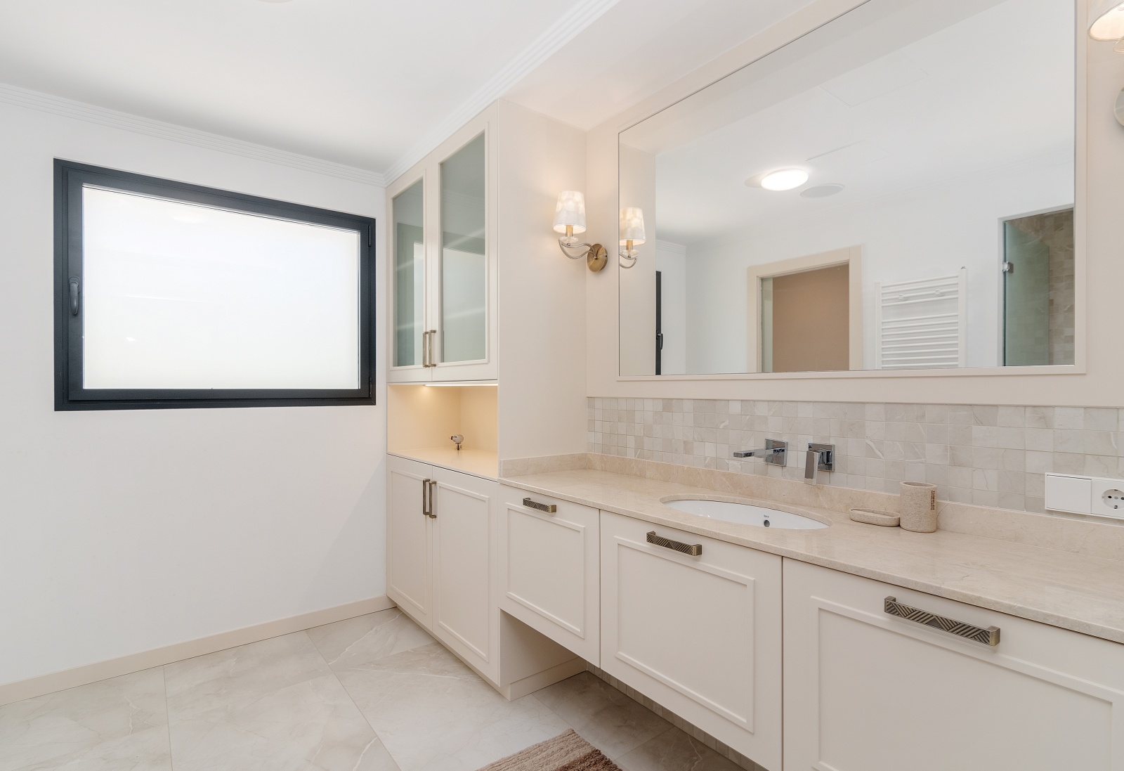 Elegant bathroom featuring a stone countertop with integrated sink and cream cabinetry. Includes glass-front display units with lighting and classic wall sconces.